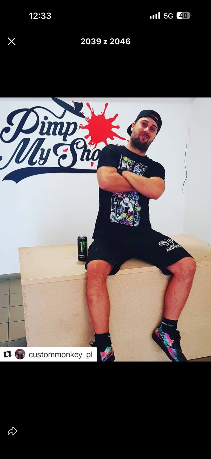 Łukasz Małpa z Custom Monkey siedzi na blacie w pracowni Pimp My Shoes, za nim logo marki
